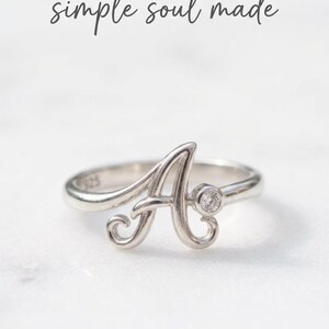 Könnte beinhalten: Ein silberfarbener Ring mit einem kursiven Buchstaben A als Mittelstück, akzentuiert durch einen kleinen runden, klaren Stein rechts neben dem Buchstaben. Die Innenseite des Rings ist mit 925 gestempelt. Der Text simple soul made steht über dem Ring.