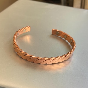 Op de afbeelding: Een koperkleurige armband met een gevlochten ontwerp. De open manchetarmband is gemaakt van gedraaide metalen strengen. De armband wordt gepresenteerd op een lichtgrijze ondergrond.