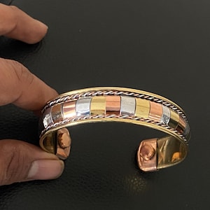 Könnte beinhalten: Ein goldfarbenes Armband mit einem mehrfarbigen rechteckigen Design. Das Armband hat kupfer-, silber- und goldfarbene Metallquadrate. Das Armband ist offen und hat einen Magnetverschluss.