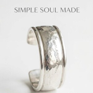 Puede incluir: Un anillo tipo brazalete de plata con textura martillada y bordes elevados. El texto SIMPLE SOUL MADE aparece impreso sobre el anillo. El anillo presenta una banda ancha con acabado pulido y una abertura lateral visible en la pieza.