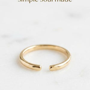 Könnte beinhalten: Ein goldfarbener offener Ring mit einer glatten, polierten Oberfläche. Der Ring weist eine kleine Lücke zwischen den beiden Enden auf. Über dem Ring ist der Text 'simple soul made' in einer dunkelbraunen Serifenschrift abgedruckt.