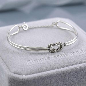 Può includere: Un braccialetto rigido color argento con un nodo al centro. Il braccialetto è realizzato in metallo sottile e delicato e ha uno stile semplice ed elegante. Il braccialetto è appoggiato su una superficie grigia.