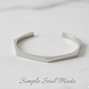 Könnte beinhalten: Ein silberfarbenes, geometrisches Armreif-Armband mit sechseckigem Design. Das offene Armband hat einen minimalistischen Stil und wird auf einer weißen Oberfläche präsentiert. Der Text "Simple Soul Made" steht darunter.