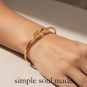 May include: A gold-coloured arrow bracelet. The arrow design features a detailed feather fletching and a pointed arrowhead. The bracelet is worn on a wrist. The words "simple soul made" are at the bottom of the image.