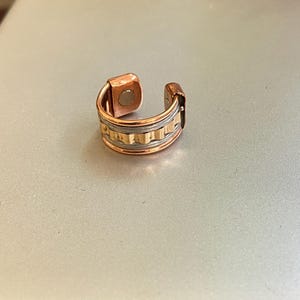 Pode incluir: Um ear cuff cor de cobre com fecho magnético. O cuff apresenta uma faixa decorativa com linhas horizontais e verticais. O ear cuff foi concebido para ser usado na orelha.