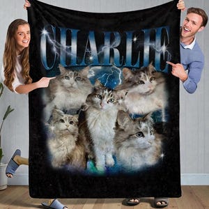 May include: Black fleece blanket featuring a collage of fluffy cats with the name "CHARLIE" in blue, stylized text with lightning bolts. The cats have gray and white fur. The blanket is held by two people.