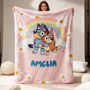 Op de afbeelding: Een roze fleece deken met een cartoonafbeelding van twee honden die elkaar omhelzen, een regenboog en madeliefjes. De naam "AMELIA" is in blauw gedrukt. De deken wordt door een persoon vastgehouden.
