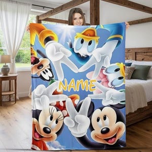 Puede incluir: Una manta azul con los personajes de dibujos animados Mickey Mouse, Minnie Mouse, Pato Donald y Goofy. La manta tiene un fondo azul claro con la palabra "NAME" en amarillo. Las manos de los personajes hacen signos de paz.