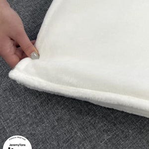 May include: Close-up of a white fleece blanket with a soft, plush texture. The blanket's edge is visible, showing a neatly sewn border. The blanket is on a gray surface. The text "Fleece Blanket" and "JeremyTans" is visible.