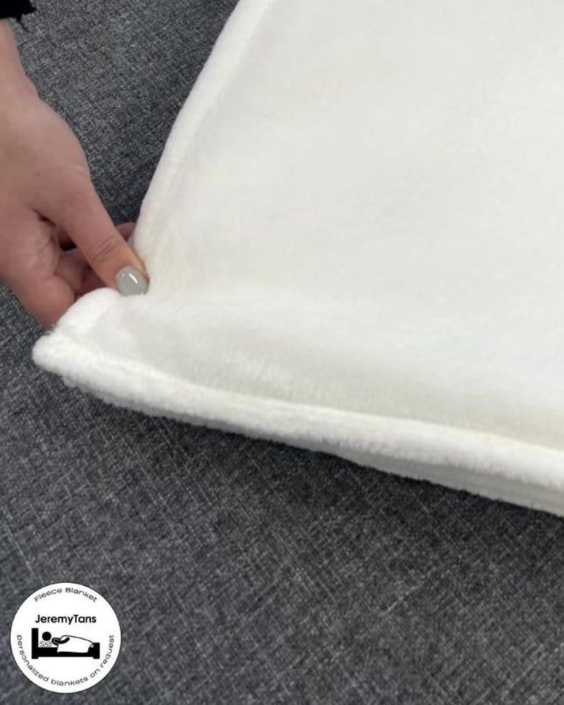 May include: Close-up of a white fleece blanket with a soft, plush texture. The blanket's edge is visible, showing a neatly sewn border. The blanket is on a gray surface. The text "Fleece Blanket" and "JeremyTans" is visible.