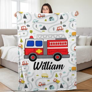 Könnte beinhalten: Weiße Decke mit einem farbenfrohen Cartoon-Design mit einem roten Feuerwehrauto, Straßen, Bäumen und Häusern. Der Name "William" ist in schwarzer Schrift darunter gedruckt. Die Decke wird von einer Person gehalten.