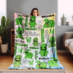 May include: A personalized blanket featuring a green cartoon character with the name "William" in various fonts. The design includes spiderwebs and the letter "W" on a pastel background. The blanket is held up by a person.
