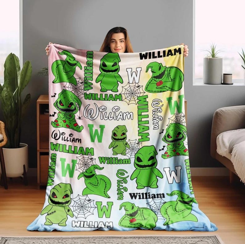 May include: A personalized blanket featuring a green cartoon character with the name "William" in various fonts. The design includes spiderwebs and the letter "W" on a pastel background. The blanket is held up by a person.