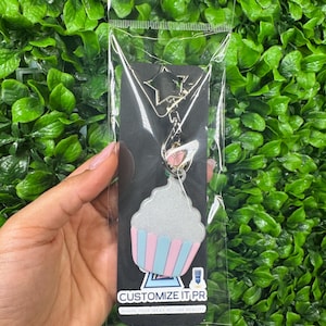 May include: A cupcake-shaped keychain with a silver glitter top and pink and blue striped base. The keychain is attached to a star-shaped charm and a metal chain. The packaging includes the text "CUSTOMIZE IT.PR".
