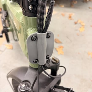 May include: Close-up of an olive green electric bicycle frame with black cables and a gray cable holder. The frame is secured with black bolts. The front wheel assembly is visible.