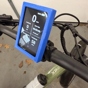May include: A blue digital display mounted on a bicycle handlebar, showing speed, trip time, and other metrics. The display is in a blue frame. A water bottle is visible below the handlebar. The bicycle frame is green.