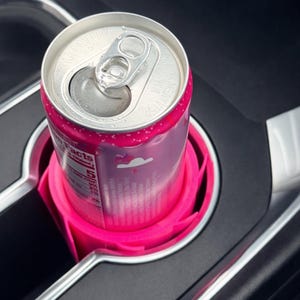 May include: A pink beverage can in a bright pink car cup holder. The can is silver with pink accents and a pull tab. The cup holder is designed to fit snugly in a car's console.