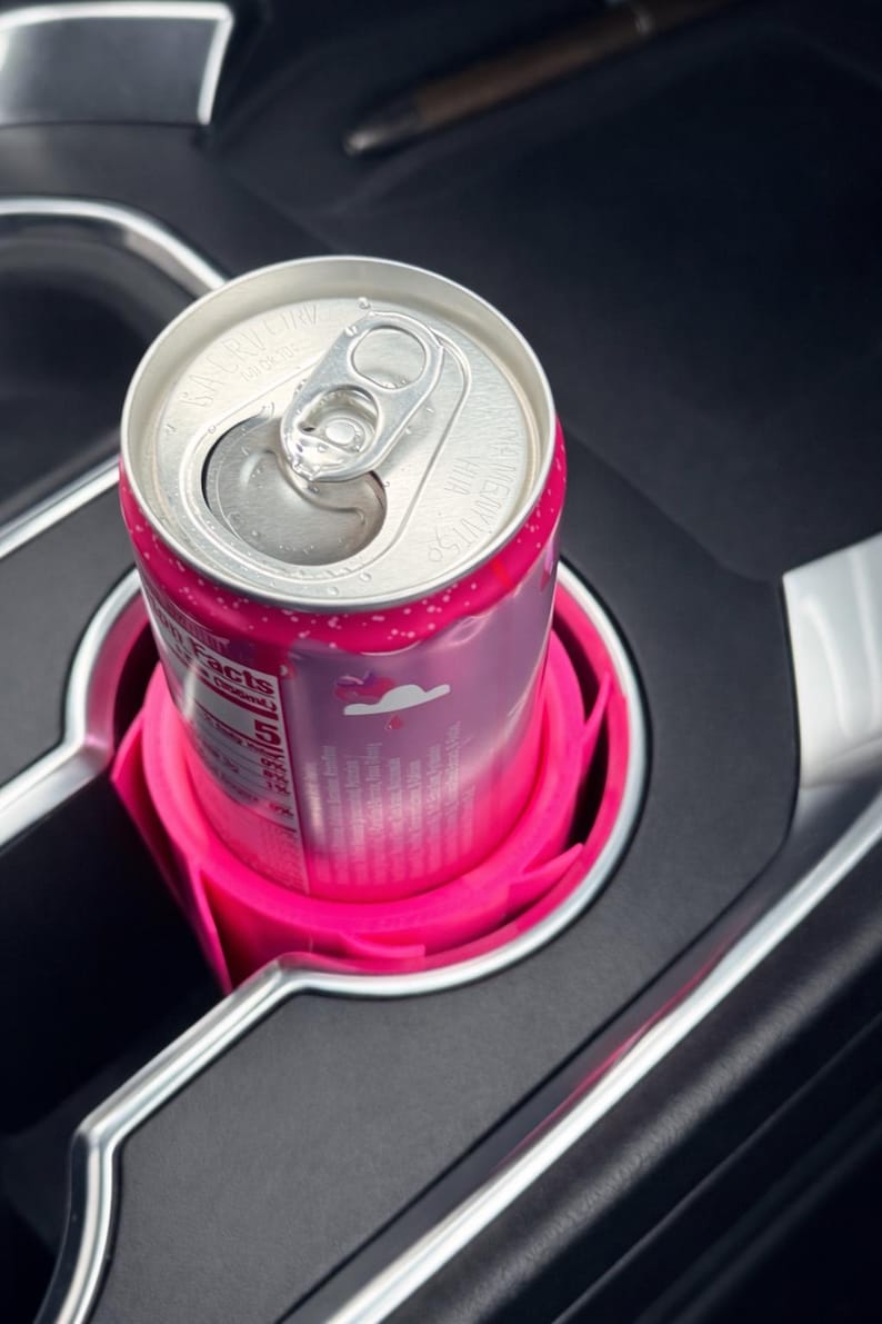 May include: A pink beverage can in a bright pink car cup holder. The can is silver with pink accents and a pull tab. The cup holder is designed to fit snugly in a car's console.