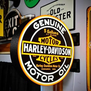 May include: A round, illuminated Harley-Davidson Motor Oil sign with a black, yellow, and white color scheme. The text reads Genuine, 5 Gallons U.S. Measure, Motor Harley-Davidson Cycles, and Harley-Davidson Motor Co. Milwaukee, Wis. mounted on a wall.
