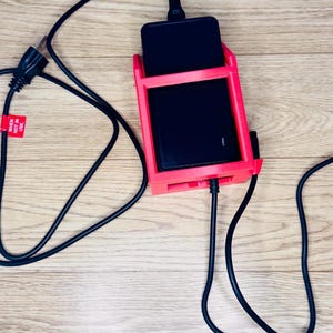 May include: A red phone charging stand with a black phone inside, connected to a black charging cable and power cord. The power cord has a label that reads "WHEN NOT IN USE".