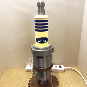May include: A unique lamp designed to resemble a large spark plug. The lamp features a silver top, a yellow body with blue stripes and the Ford logo, and a brown gear-shaped base. The lamp is plugged into a power outlet.