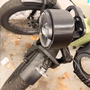May include: A black circular headlight mounted on the front of a light green electric bicycle. The headlight features a clear lens and a black housing. Below the headlight, a small clear turn signal light is attached to the front fork assembly.