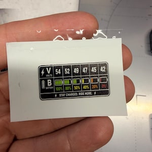May include: A rectangular black sticker with white text and graphics displaying a battery voltage chart. The chart includes columns for 54, 52, 49, 47, 45, and 42 volts, corresponding battery icons with percentage levels, and the text STAY CHARGED. RIDE MORE.