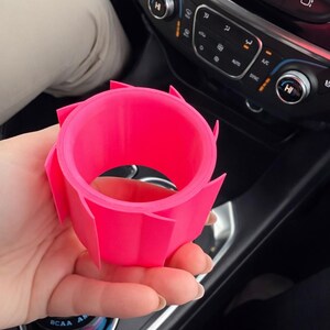 May include: A bright pink, 3D-printed car cup holder. The holder has a circular interior and a geometric, petal-like exterior design. The cup holder is held in a person's hand, with a car dashboard visible in the background.