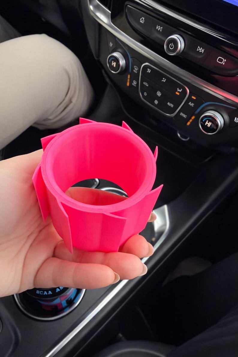 May include: A bright pink, 3D-printed car cup holder. The holder has a circular interior and a geometric, petal-like exterior design. The cup holder is held in a person's hand, with a car dashboard visible in the background.