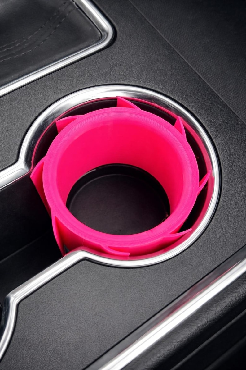 May include: A bright pink car cup holder insert, designed to fit inside a vehicle's existing cup holder. The insert has a unique, angular design with several protruding tabs. The surrounding area is black with a silver trim.