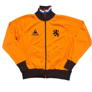 May include: Orange track jacket with black accents and a full zipper. Features a black lion emblem and the words "le coq sportif". The collar has red, white, and blue stripes. The cuffs and bottom hem are black.