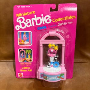 Op de afbeelding: Een roze Barbie verzamelobject uit 1989, met een miniatuur Barbie figuur in een roze outfit op een geblokte basis. De verpakking is felroze met de woorden "Miniature Barbie Collectibles" en "Cool Times Barbie 1989".