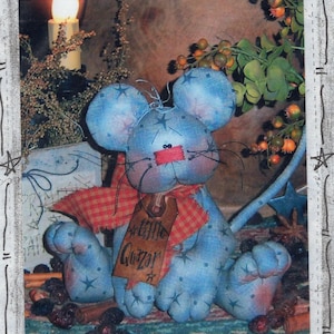 May include: A handmade blue mouse doll with star accents, a red plaid scarf, and a tag that reads "Little Quazar". The doll has large ears and a long tail. The background includes a candle and greenery.