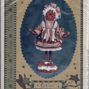 May include: A Christmas-themed craft pattern featuring a gingerbread doll named "Gretchen." The doll wears a dress with a white apron and stands on a cake base. The image has a blue oval background with stars and the text "Sparkles 'N' Spirit."