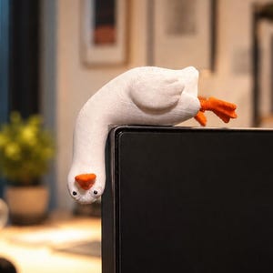 May include: A white plush goose decoratively draped over a black computer monitor. The goose has an orange beak and feet. The background is blurred, but includes a desk, window, and artwork.