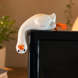 May include: A white goose figurine with an orange beak and feet, designed to perch on the top corner of a computer monitor. The text Goose Monitor Buddy and Little goose. Big personality. appear above the figurine, with icons below.