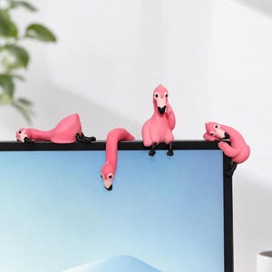 May include: Four small pink flamingo figurines with black beaks and feet, designed to perch on the top edge of a computer monitor. Each flamingo is in a different pose, including one lying down, one hanging over the edge, one sitting, and one resting.