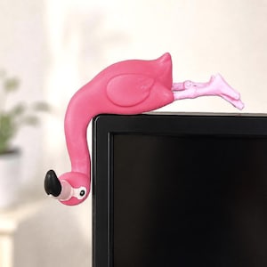 May include: A bright pink flamingo figurine designed to perch on the top corner of a monitor. The flamingo has a long curved neck, a black-tipped beak, and its legs are extended horizontally along the top edge of the black screen frame.