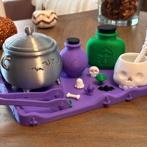 Può includere: Un vassoio viola contenente un calderone argentato con intagli a forma di pipistrello, una boccetta viola con una luna, una boccetta verde con funghi, una tazza a forma di teschio bianco con cucchiaio scheletrico e piccole statuine.