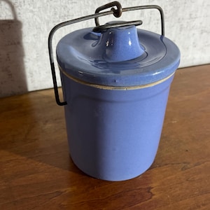 May include: A small, blue ceramic jar with a lid and a metal handle. The jar has a cylindrical shape and a slightly textured surface. The lid fits snugly on top, and the handle is attached to the sides. The jar is sitting on a wooden surface.