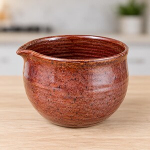 May include: A round ceramic pouring bowl with a glossy, mottled reddish-brown glaze. The exterior features subtle horizontal ridges along the sides, and a small, integrated spout is positioned on the upper left rim for controlled pouring.