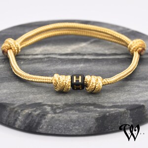 Könnte beinhalten: Ein goldfarbenes Seilarmband mit einer schwarzen Würfelperle mit dem Buchstaben "H". Das Armband ist an jedem Ende und in der Mitte geknotet, mit einem doppelsträngigen Design. Das Armband wird auf einer grauen, strukturierten Oberfläche präsentiert.