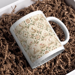 Może przedstawiać: Biały ceramiczny kubek z powtarzającym się wzorem przedstawiającym kr&oacute;liki, grzyby i motywy kwiatowe w stonowanych odcieniach zieleni, brązu i kremu. Wz&oacute;r jest ułożony w siatkę z naprzemiennymi kwadratowymi i okrągłymi elementami dekoracyjnymi.