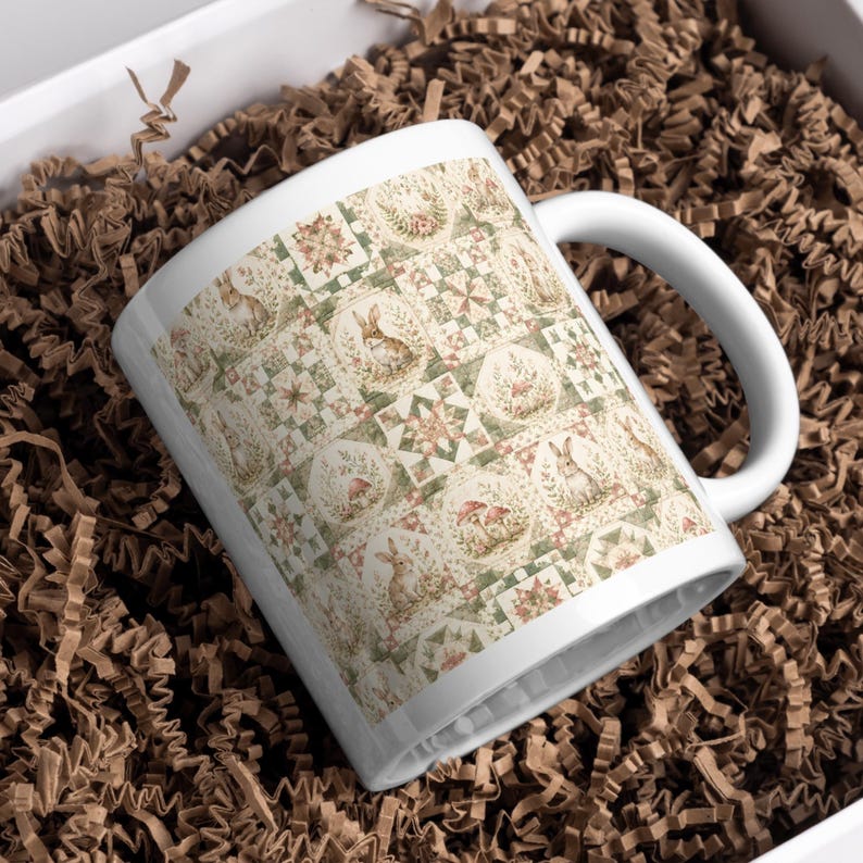 Może przedstawiać: Biały ceramiczny kubek z powtarzającym się wzorem przedstawiającym kr&oacute;liki, grzyby i motywy kwiatowe w stonowanych odcieniach zieleni, brązu i kremu. Wz&oacute;r jest ułożony w siatkę z naprzemiennymi kwadratowymi i okrągłymi elementami dekoracyjnymi.