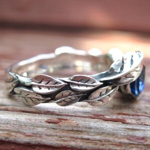 Puede incluir: Un anillo de plata con una piedra preciosa azul engastada en un dise&ntilde;o de hojas. La banda est&aacute; decorada con una fila de hojas.