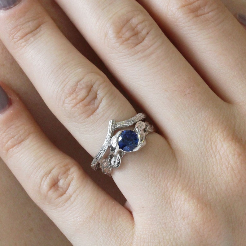 Puede incluir: Un anillo de plata con una piedra preciosa de zafiro azul engastada en un dise&ntilde;o de rama. El anillo se lleva en un dedo con esmalte de u&ntilde;as transparente.