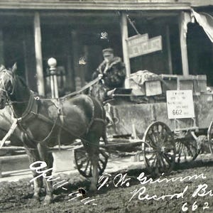 May include: A black and white photograph of a horse-drawn cart. Two horses are hitched to the cart, which is loaded with boxes. A person is seated in the cart. A sign reads "Just Received" and another sign reads "662". The text "J.W. Grimm with Record Breaker hall 662 doz. eggs" is written on the image.