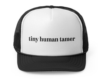 May include: A black and white trucker hat with the text "tiny human tamer" in black. The hat features a white front panel, a black brim, and a mesh back.
