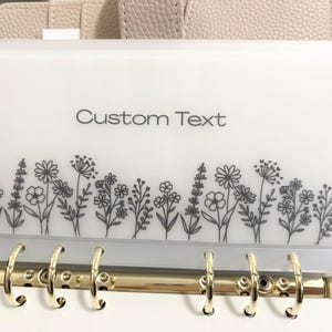 May include: A clear, white binder pocket with gold rings, featuring a floral design along the bottom and the words "Custom Text" at the top. The floral design is a series of black line drawings of various flowers and plants.