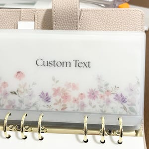May include: A clear plastic binder pocket with floral accents in pink and purple, featuring the text "Custom Text". The pocket is part of a beige binder with gold rings.